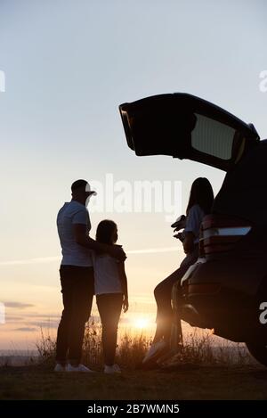 Silhouette der dreiköpfigen Familie, die den Sonnenuntergang auf dem Hügel außerhalb der Stadt genießt, Frau sitzt im Kofferraum, Vater steht auf und umarmt seine Tochter Stockfoto