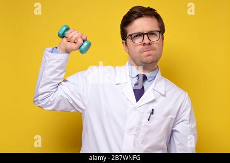 Ein reifer kaukasischer Mannarzt, der Hanteln hält und Ratschläge zu einem gesunden Lebensstil gibt. Sport- und Medizinkonzept an gelber Wand. Stockfoto