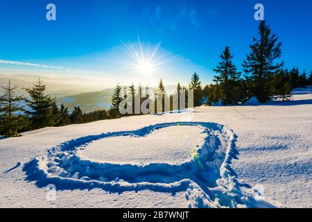 Herz trampelte auf dem Schnee mit Füßen in einer Schneeverwehung auf einem Hang mit herrlichem Blick auf den Nadelwald und die Bergketten auf einem sonnigen Frost Stockfoto