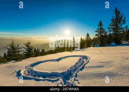 Herz trampelte auf dem Schnee mit Füßen in einer Schneeverwehung auf einem Hang mit herrlichem Blick auf den Nadelwald und die Bergketten auf einem sonnigen Frost Stockfoto