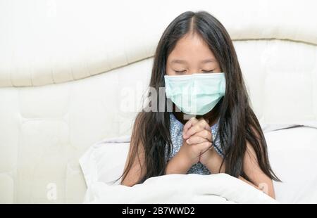 Kleines mädchen aus asien, das eine hygienische Maske zum Schutz der pm2.5 und Covid-19 trägt, betete sie auf dem Bett für einen neuen Tag Freiheit für den wuhan Corona Virus. Stockfoto