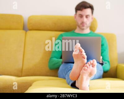 Junger kaukasischer Mann, der seit dem Ausbruch des Coronavirus in Europa von zu Hause aus auf seinem Computer auf einem Sofa sitzt. Stockfoto