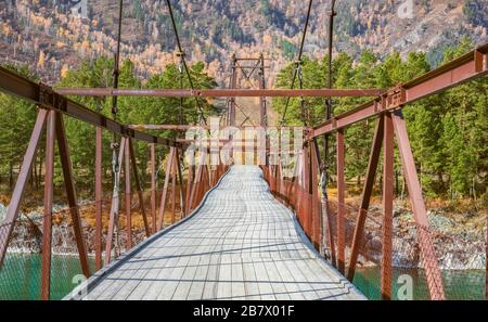 Hängebrücke, rostige Brücke umgeben von Bäumen und Bergen. Reduzierung der Perspektive einer Fußgängerbrücke mit Metallfederung über das türkisfarbene Katu Stockfoto