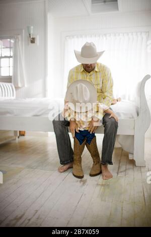 Kleiner Junge in großen Hut- und Cowboystiefeln, der sich neben einem Bett in einem Haus an seinen Vater lehnt. Stockfoto
