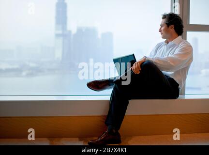 Der Geschäftsmann, der seinen Laptop benutzt, sitzt auf einem Fenstervorsprung, der über die Skyline der Stadt blickt. Stockfoto