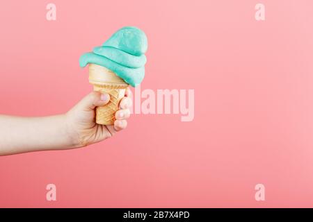 Eis in der Hand eines Kindes schmilzt auf einem rosafarbenen Hintergrund. Der Waffelkegel mit blauem Eis schmilzt. Stockfoto