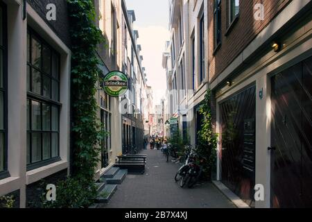 Blick auf die Menschen, die auf einer der engen Straßen in Amsterdam spazieren. Berühmte lokale Bierschilder und historische Gebäude sind in Sicht. Es ist ein sonniges su Stockfoto