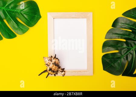 Tropischer Mock mit weißem Bilderrahmen und Palmblättern auf farbenfrohem gelbem Hintergrund. Fotorahmen mit leerem Platz für Text oder Werbung mit Muscheln auf gelbem Hintergrund. Stockfoto