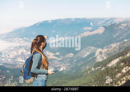 Junges, wunderschönes Mädchen reist allein im Frühjahr oder Herbst in die Berge, schaut mit dem Telefon in die Ferne und genießt Natur, Felsen und grüne Wälder, Blick auf die Landschaft, einen Rucksack dahinter und Sportswear, Freiheit und Leichtigkeit Stockfoto