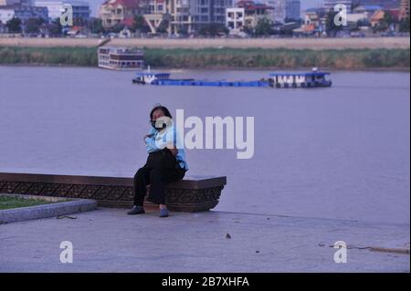 Eine ältere kambodische Frau in einer Gesichtsmaske sitzt während der Coronavirus-Pandemie allein am menschenleeren Flussufer. Phnom Penh, Kambodscha. © Kraig Lieb Stockfoto