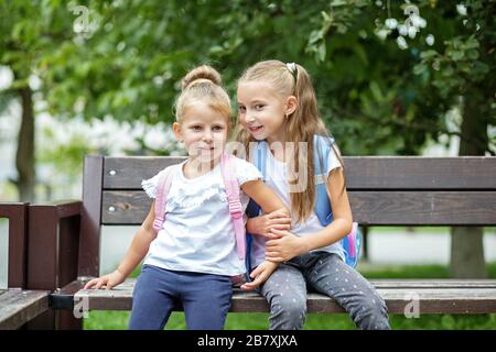 Zwei kleine Kinder klatschen auf einer Bank. Mädchen mit Rucksäcken. Das Konzept geht zurück auf Schule, Familie, Freundschaft und Kindheit. Stockfoto