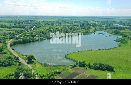 Das blaue Wasser im See wird vom Himmel geschossen. Grüne im Dorf. Viele Pflanzen und Bäume. Luftansicht von oben auf ein grünes Feld und einen kleinen Schweller Stockfoto