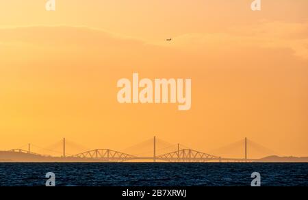 Firth of Forth, Schottland, Großbritannien. März 2020. Wetter in Großbritannien: Ein farbenfroher orangefarbener Himmel bei Sonnenuntergang über dem markanten Umriss der drei vierten Brücken mit einem Flugzeug am Himmel beim Abstieg zum Flughafen Edinburgh, von East Lothian aus gesehen Stockfoto