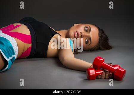 Müde hübsche junge Sportlerin, die rote Hanteln in den Händen hält, während sie auf dem grauen Boden liegt. Konzept der Ermüdung aus dem Training. Copyspace Stockfoto