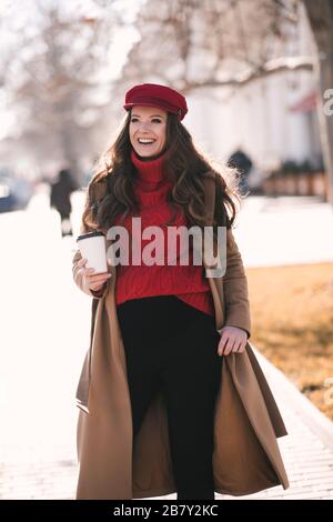 Lächelnde schwangere Frau, die auf Straßen spazieren ging und Kaffee über dem Hintergrund der Stadt trinkt. Frühlingssaison. Stockfoto