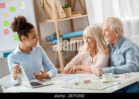 Afrikanische Abstiegsfrau Reisebüro im Büro, die Firmenwebsite auf dem Laptop zu älteren Paaren zeigt, die neugierig lächelnd fröhlich aussehen Stockfoto