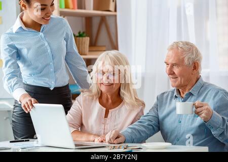 Afrikanische Abstiegsfrau Reisebüro im Büro in der Nähe eines älteren Paares, das am Tisch sitzt und heißen Kaffee trinkt, der im Internet auf dem Laptop lächelnd chee surft Stockfoto