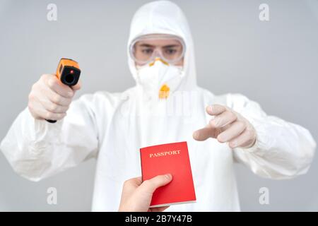Coronavirus-Konzept. Arzt in Gefahrgut Anzug und Atemschutzmaske auf grau isoliert stehend mit Pass der chinesischen Person hält Infrarotkamera Stockfoto