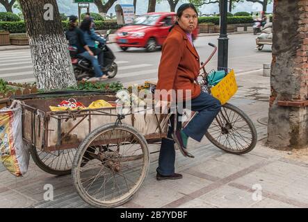 Guilin, China - 11. Mai 2010: Stadtzentrum. Frau steht mit Transportdreirad mit großer Ladeabteilung auf der Straßenseite. Stockfoto