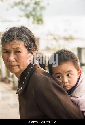 Guilin, China - 11. Mai 2010: Stadtzentrum. Nahaufnahme der Großmutter in brauner Garbe, die den jungen Jungen auf dem Rücken trägt. Stockfoto