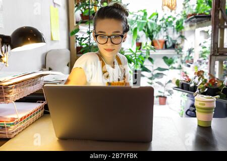 Junge Gärtnerin in Brille mit Laptop, kommuniziert im Internet mit dem Kunden im Garten, wiederverwendbarer Kaffee-/Teebecher auf dem Tisch. Zuhause arbeiten Stockfoto
