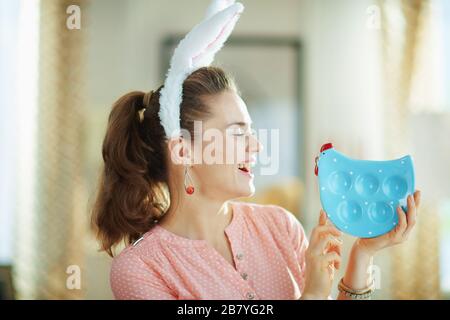 Lächelnde, elegante 40 Jahre alte Frau in einer rosafarbenen Bluse und osterbrötchen im modernen Zuhause am sonnigen Frühlingstag mit Blick auf die blaue Hühnerform Stockfoto