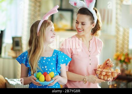 Fröhliche moderne Mutter und Tochter in den österlichen Hunnenohren mit blauem Hähnchenschild mit bunten Eiern und Korb mit Bauerneiern im modernen hou Stockfoto