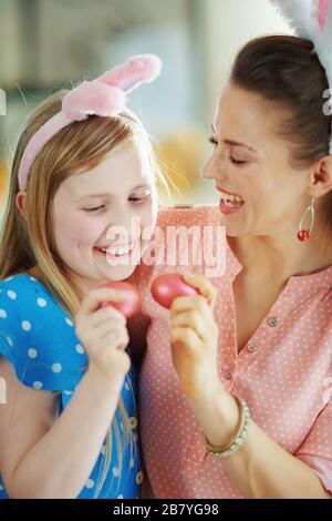 Glückliche junge Mutter und Kind in den osterbrunnenohren, die an einem sonnigen Frühlingstag im modernen Zuhause Eierklopfen mit roten ostereiern haben. Stockfoto