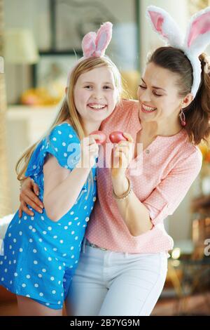 Fröhliche elegante Mutter und Tochter in den österlichen Hünenohren, die am sonnigen Frühlingstag im modernen Zuhause Eierklopfen mit roten ostereiern haben. Stockfoto