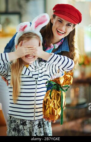 Die Mutter, die der Tochter im modernen Wohnzimmer am sonnigen Frühlingstag in osterhasen ein großes osterei in Goldfolie präsentiert. Stockfoto