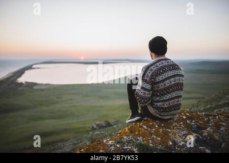 Junger Mann im gestreiften Pullover sitzt auf Hügel während des Sonnenuntergangs Stockfoto