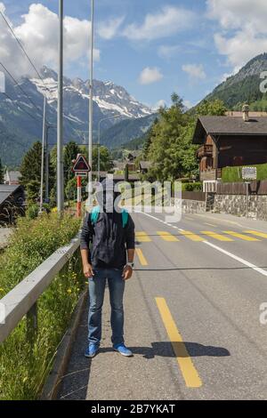 Der junge Mann verwendet eine Nackengage, braga, die wie eine medizinische Gesichtsmaske aussieht. Warmer sonniger Tag, blauer Himmel mit weißen Wolken. Schweizer Alpen im Hintergrund. Coron Stockfoto