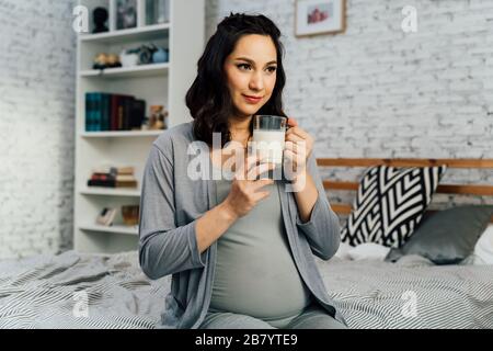 Froh schwangere Mutter mit gemischten Rassen sitzt auf dem Bett und trinkt Milch, während sie in einem gemütlichen Schlafzimmer zu Hause ruht Stockfoto