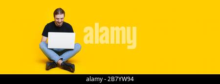 Bärtiger kaukasischer Mann mit langen Haaren sitzt auf einer gelben Wand mit freiem Platz und arbeitet am pc Stockfoto