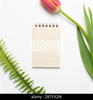 Leerer Notizblock mit Tulpenblume auf weißem Hintergrund. Blumendesign, flacher Lay, Draufsicht, Kopierbereich Stockfoto