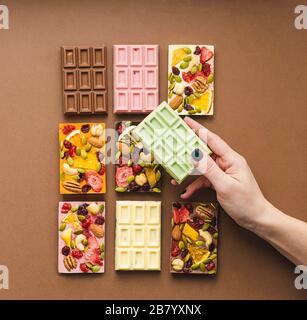 Frau mit heller Maniküre hält in der Hand Schokolade verschiedener Farben und farbige Füllungen mit Scheiben von Fruchtnüssen. Quadratischer Rahmen Stockfoto