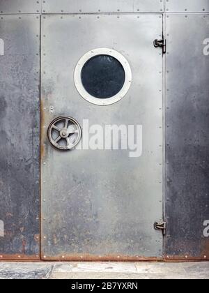 Tür mit Porthole. Metalltür mit runder Scheibe auf rostendem Wandhintergrund aus Metall Stockfoto