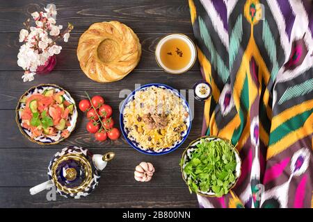 Nationaler usbekischer Pilaf mit Fleisch, Achichuk-Salat aus Tomaten, Gurke, Zwiebel in Platte mit traditionellem Muster, Cilantro, Kirschtomaten, Knoblauch, Brot Stockfoto