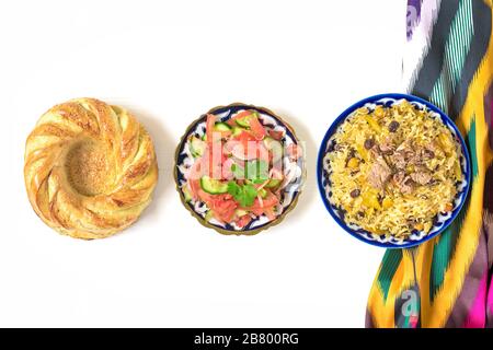 Nationaler usbekischer Pilaf mit Fleisch, Achichuk-Salat aus Tomaten, Gurke, Zwiebel in Platte mit traditionellem Muster, Cilantro, Kirschtomaten, Knoblauch, Brot Stockfoto