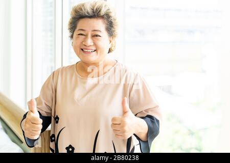 Porträt des fröhlichen und lächelnd älteren älteren Erwachsenen Frau neben dem Fenster im häuslichen Wohnzimmer stehend - Gesundung und Rehabilitationskonzept Stockfoto