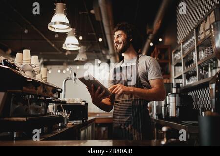 Fröhlicher junger kaukasischer Café-Besitzer, der Schürze mit digitalem Tablet trägt Stockfoto