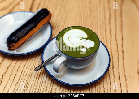 Matcha grün in einem weißen Becher auf einem Holztisch in einem Café neben einem süßen Dessert. Schokolade eclair zum Frühstück. Stockfoto