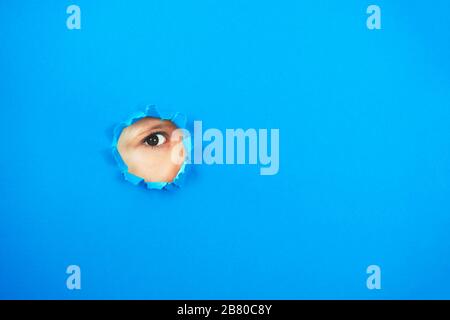 Das Auge des Kindes sieht durch das Loch in blauem Papier. Spionieren Sie die Augen durch das Loch. Kopierbereich. Stockfoto