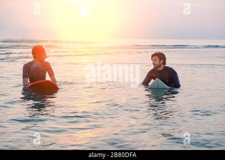 Zwei Männer in Wetsuits mit Surfbrett an einem sonnigen Tag im Wasser - Strandsurfer sprechen und lachen, warten auf Wellen bei Sonnenaufgang - Extreme Sports A Stockfoto