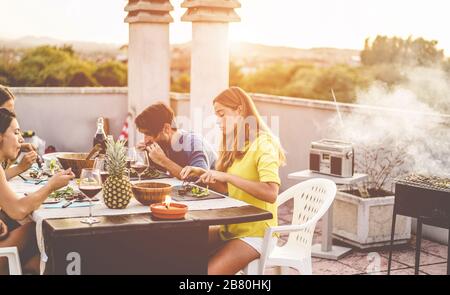 Junge Freunde, die bei Sonnenuntergang auf der Dachterrasse grillen - fröhliche Leute, die ein grillabendessen im Freien machen, Fleisch kochen und Wein trinken - konzentrieren sich auf die rechte Seite Stockfoto