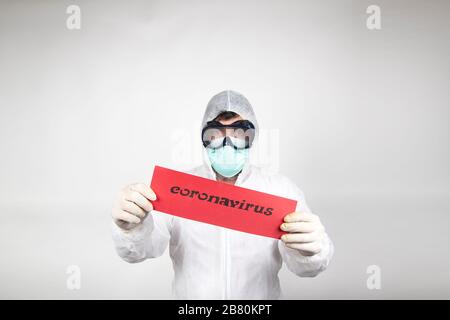 Mann mit chirurgischer Maske, Brille, weißem Schutzanzug und rotem Schild mit dem Wort Coronavirus isoliert im Studio auf weißem Hintergrund. Neue pandemische Epi Stockfoto