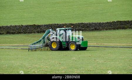 Farmer beschäftigt sich mit der Arbeit auf der Farm und fährt mit dem grünen Traktor, der Gülle mit nachlaufenden Schläuchen verbreitet, um Ackerland Weidefeld zu befruchten - Yorkshire, England, Großbritannien. Stockfoto