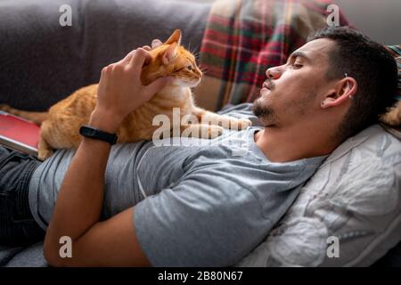 Tabby-Katze liegt auf der Brust eines jungen Mannes, der auf dem Sofa unter dem Fenster liegt Stockfoto