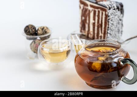 Blitzender schwarzer Tee in einer Glaskanne. Auf einer hellen Oberfläche. In der Nähe liegen Teebriketts mit Blättern und Blumen. Im Hintergrund ein Kuchen. Kopierbereich Stockfoto