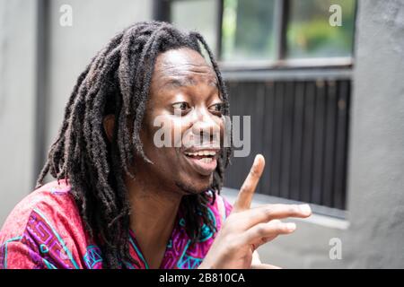 Junger schwarzer Mann mit Dreadlocks und einem farbenfrohen Hemd mit einem fröhlichen Gesichtsausdruck beim Anheben eines Fingers Stockfoto